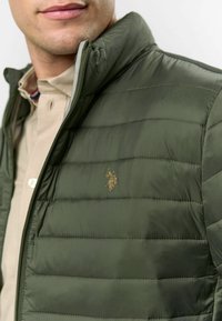 Zelená nafukovacia bunda so zipsom, horizontálnym prešívaním a zlatým logom na hrudi. Nosená cez svetlú košeľu.