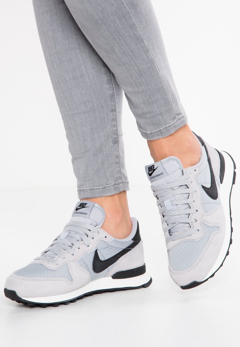 Personne portant des baskets Nike gris clair avec le logo Swoosh noir et un pantalon gris, debout sur une surface blanche.