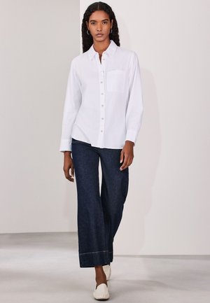 Hosszú, göndör hajú nő fehér gombos ingben, sötét, bő szárú farmerral és fehér lábbeliben, sima háttér előtt áll.