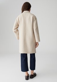 Vrouw met lang haar in een beige jas tot de knie, cropped donkerblauwe jeans en donkeren bruine loafers, staand tegen een effen achtergrond.