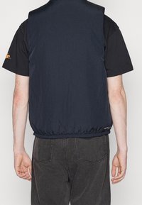 Marinblå ärmlös väst gjord av lätt material med texturerad yta, med elastisk nederkant och logotypdetalj längst ner.