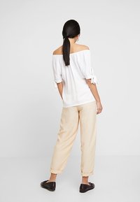 Top bianco off-the-shoulder con maniche corte e dettagli in stile cravatta, abbinato a pantaloni beige chiaro e larghi e scarpe nere.