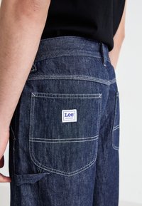 Denim workwear pants featuring a dark blue color, visible stitching, multiple pockets, and a Lee® label on the back pocket.