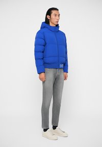 Kék párnázott dzseki kapucnival, cipzáros elülső résszel és logóval ellátott folttal, szürke farmerral és fehér sneakerrel párosítva. Simább szövet textúra.