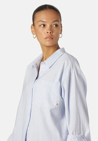 Camicia azzurro chiaro con bottoni, dotata di tasca sul petto, in tessuto morbido, vestibilità rilassata e maniche arrotolate. Presenta un'accenno di logo discreto sulla tasca.