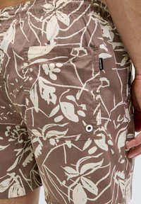 Pantaloni corti marroni con un motivo floreale bianco, dotati di un elastico in vita e di una tasca a toppa con dettaglio in occhio di metallo.