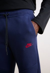 Marineblå sweatpants i blødt stof med en sort snoretræk og et rødt Nike-logo på venstre lår. Designet har tætsiddende ben.