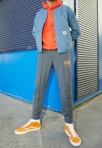 Vadderad ljusblå jacka, orange hoodie, grå Nike sweatpants med guldaccenter, samt orange Vans-skor med vita sulor, framför en blå bakgrund.