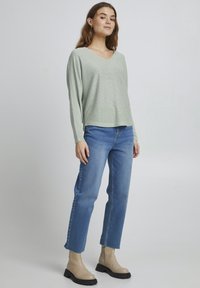 Hellgrünes, strukturiertes Langarmshirt mit V-Ausschnitt, kombiniert mit mittelblauen, geraden Jeans und beigen Ankle-Boots mit stabilen Sohlen.