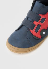 Kék velúr cipő piros és fekete részletekkel, lekerekített orral és két rugalmas pánttal a záródásért, bézs gumitalpon pihen.