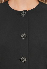 Černý látkový blazer s třemi ozdobnými knoflíky ve tvaru květin se rhinestony. Design má kulatý výstřih a hladkou texturu.