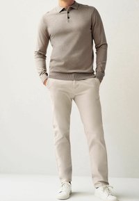 Pullover a maglia color marrone chiaro con colletto e pattina con tre bottoni, abbinato a pantaloni beige slim-fit e sneakers bianche.