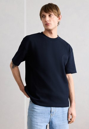 Νέος άνδρας με ανοιχτό καστανά μαλλιά, που φοράει ένα φαρδύ σκούρο μπλε κοντομάνικο πουκάμισο και ανοιχτό μπλε τζιν, κοιτάζοντας στο πλάι με το ένα χέρι στην τσέπη.