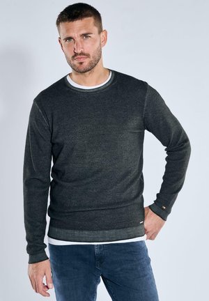 Man with short hair and beard wearing dark gray sweater over white shirt and blue jeans, standing with one hand on hip.