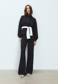 Pull noir en tricot avec un col montant et fermeture éclair, associé à un pantalon évasé assorti. Une ceinture blanche souligne la taille. Des chaussures à bout pointu présentent un imprimé serpent.