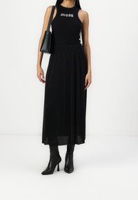 Top noir sans manches avec le texte "MUSE", associé à une jupe maxi plissée noire. Des bottes à talons hauts noires complètent l'ensemble.