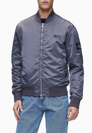 Mann in blauer Bomberjacke mit Frontreißverschluss, gerippten Bündchen an Ärmeln und Saum, kleinem Logo auf Brust und Ärmel, trägt hellblaue Jeans.