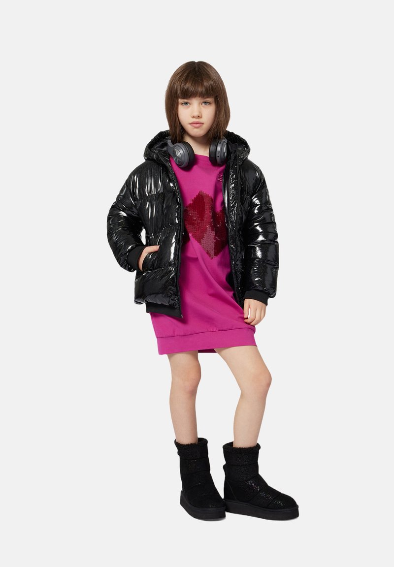 Doudoune noire, texture brillante, portée sur un sweat-shirt rose avec un design de cœur en sequins. Associée à des bottes en fourrure noires et des écouteurs.