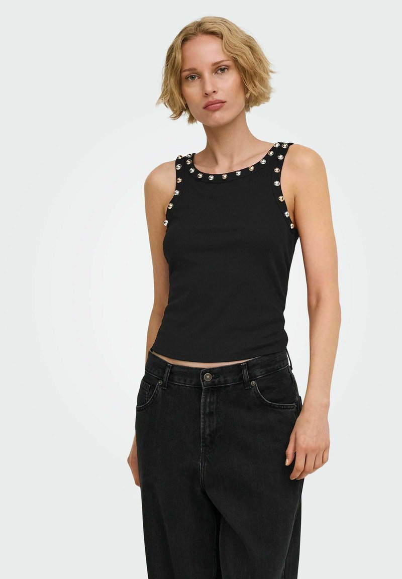Femme aux cheveux blonds courts, portant un haut sans manches noir avec des clous argentés et un jean noir taille haute, debout devant un fond uni.