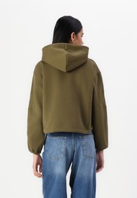 Κοντό ελαιοπράσινο φούτερ με μεγάλη κουκούλα, υφασμάτινα μανίκια και ραμμένο λογότυπο στο αριστερό μανίκι, σε συνδυασμό με μπλε τζιν παντελόνι.