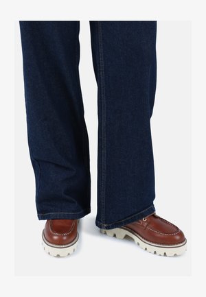 Rjave usnjene čevlje z belimi gumijastimi podplati, v kombinaciji s temno modrimi širokimi hlačami. Vidna šivna podrobnost na robu hlač.