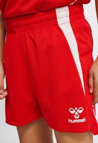 Pantaloncini sportivi rossi con strisce laterali bianche e logo Hummel bianco sulla parte inferiore della coscia sinistra, indossati da una persona con le mani rilassate lungo i fianchi.