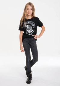 Schwarzes Kurzarm-T-Shirt mit einem weißen Hogwarts-Wappen, kombiniert mit dunkelgrauen Skinny-Jeans und schwarzen ankle boots.