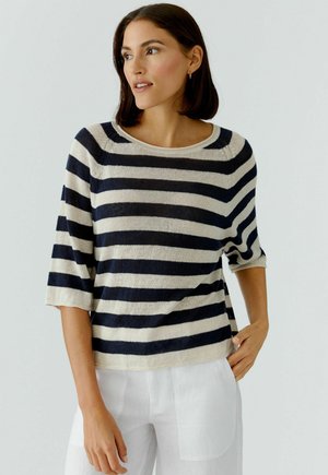 Vrouw met donker haar draagt een marineblauw-wit gestreept shirt met driekwart mouwen en een witte broek, staand met één hand in haar zak.