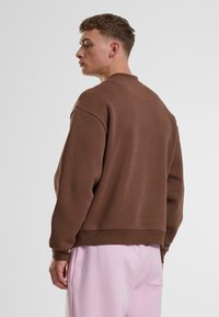 Sudadera marrón con cuello redondo y costuras de hombro caídas. Textura suave, ajuste relajado y dobladillo acanalado. Combinada con pantalones rosa claro.