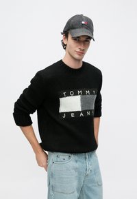 Μαύρο πλεκτό πουλόβερ με λογότυπο «TOMMY JEANS», με σχέδιο σε γκρι και ανοιχτό γκρι μπλοκ. Συνδυασμένο με ανοιχτόχρωμο μπλε τζιν παντελόνι και σκούφο σε σκούρο χρώμα.