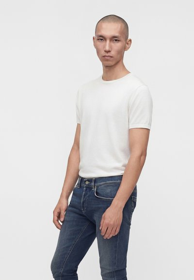 Jeune homme à la tête rasée portant un t-shirt blanc uni à manches courtes et un jean bleu sur un fond gris clair.