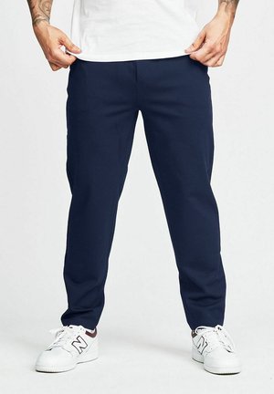Man som bär marinblå byxor, vit t-shirt och vita sneakers, står mot en enfärgad ljus bakgrund.