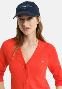 Red cardigan with a V-neck and long sleeves, featuring two buttons and a small embroidered logo. Navy cap with white embroidered text and logo.