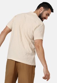 Beige kurzärmliges Hemd mit einer subtilen Musterung und Kragen; getragen mit braunen Hosen, von hinten und von der Seite sichtbar.