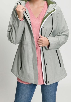 Person iført en lysegrå hættetrøje over en lyserød strikket sweater og blå jeans, der holder jakken delvist åben foran.
