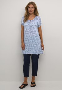 Femme portant un tunique à motifs bleu et blanc, un jean cropped bleu marine et des sandales noires à enfiler, se tenant devant un fond clair.