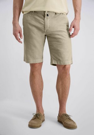 Man die beige knielange shorts en bijpassende beige casual schoenen draagt, staand op een lichtgrijze achtergrond.