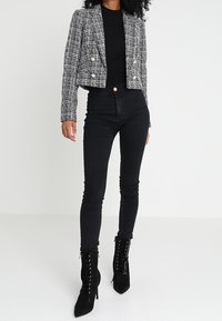Veste courte en tweed noir et blanc avec boutons dorés, associée à un jean noir taille haute et des bottines noires à lacets. Texture lisse.
