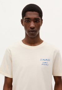 Tricou de culoare crem, cu mânecă scurtă, confecționat dintr-un material moale, având inscripționat cu text albastru scris de mână "SCALPERS SUMMER ESSENTIALS" pe piept.