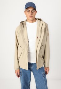 Beige vanntett jakke med hette, frontlås, to store lommer og subtil merkevare. Brukt over en hvit t-skjorte og blå jeans.