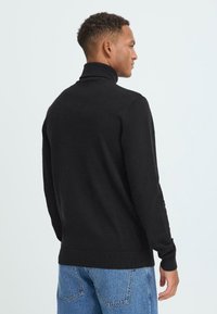 Schwarzer Strickpullover mit hohem Kragen, gerippten Bündchen und Saum. Der Rücken hat ein nahtloses Design, kombiniert mit hellblauen Jeans.