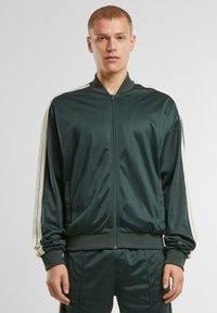 Tamnozeleni zip-up dukserica od glatkog materijala s dugim rukavima. Ima bijele bočne pruge te rebrasti ovratnik i manžete.