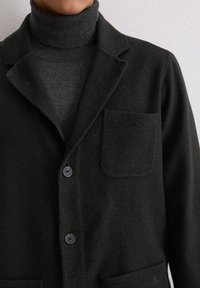 Schwarze Jacke aus Wollmischung mit Reverskragen, zwei Knöpfen, aufgesetzter Tasche und weicher Textur, getragen über einem dunkelgrauen Rollkragenpullover.