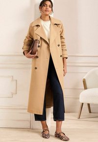 Trench coat bej cu un design cu dublă captură, clape mari și nasturi. Asortat cu blugi scurți negri și balerini cu imprimeu de leopard.