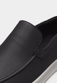 Mocassin noir texturé à enfiler avec une semelle en caoutchouc épaisse blanche, un bout arrondi et des coutures visibles sur la tige lisse.