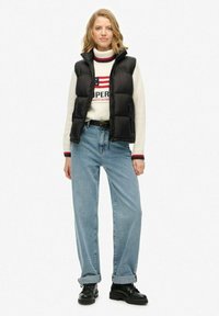 Junge Frau steht da und trägt eine schwarze Daunenweste, einen weißen Pullover mit roten und blauen Details, hellblaue hochtaillierte Jeans und schwarze Stiefel.