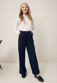 Balti marškiniai su apykakle, tamsiai mėlynos, plačiomis kojų kelnės su dviem auksiniais mygtukais ir juodi slip-on batai su kutais.