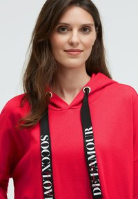 Rote Kapuzenjacke mit Schnürzug und schwarzen Textakzenten. Weicher Stoff, lockere Passform und Fronttasche im Känguru-Stil.