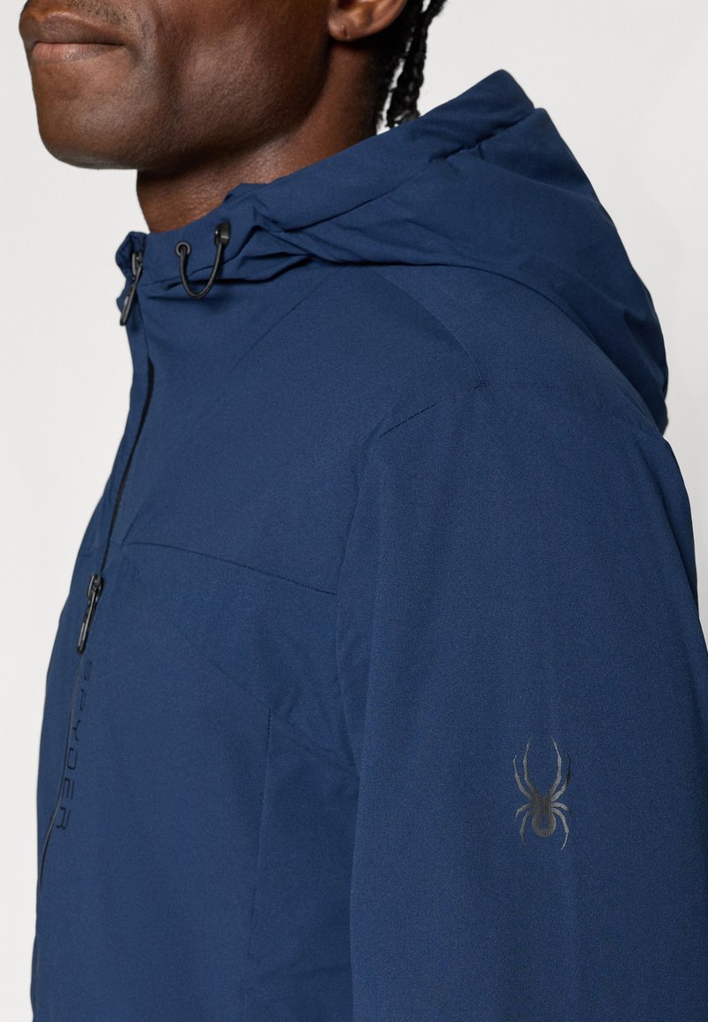 Chaqueta impermeable azul marino con capucha, tejido texturizado, bolsillo con cremallera y un logo de araña negra en el brazo izquierdo.