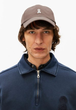 Giovane uomo con capelli castani di media lunghezza, indossa un berretto taupe con logo bianco e una giacca con zip blu navy su sfondo bianco.
